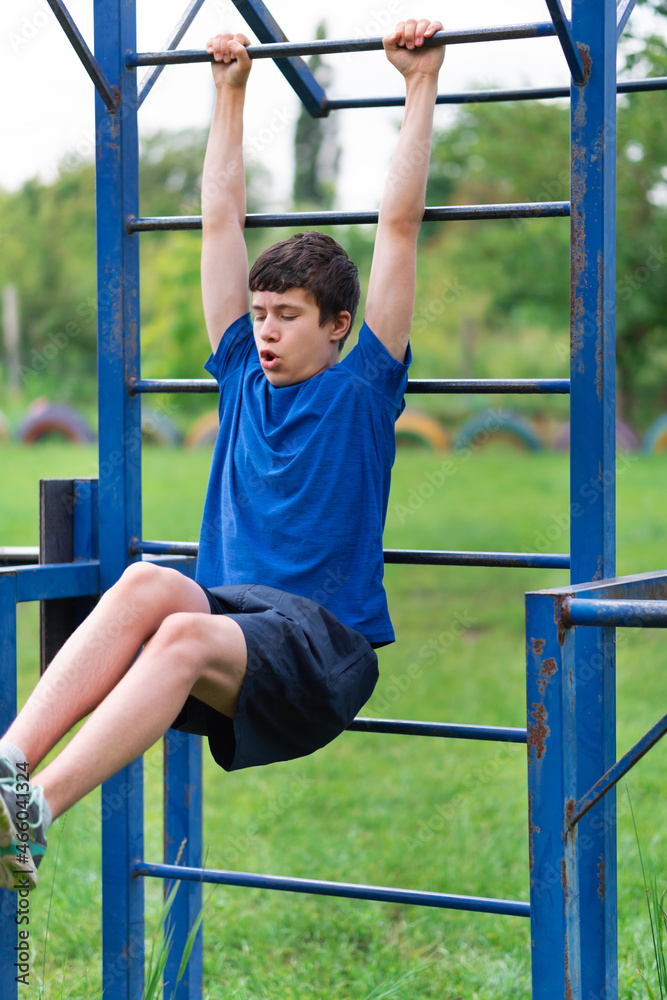 Fototapeta premium teenage boy exercising outdoors, sports ground in the yard, he hangs on the horizontal bar, raises his legs and pumps his abs, healthy lifestyle