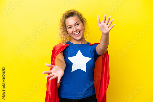 Girl with curly hair isolated on yellow background in superhero costume and doing coming gesture