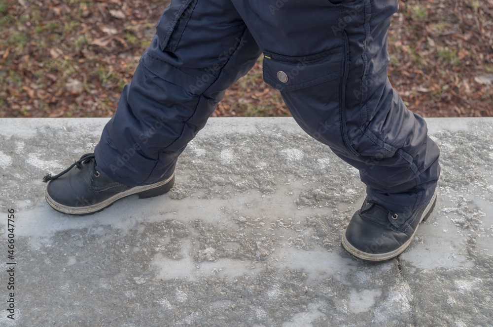 Stepping feet of a boy dressed in dark blue winter pants and black boots, walking on a snow-covered icy concrete parapet rising against a blurred background of fallen withered brown foliage.