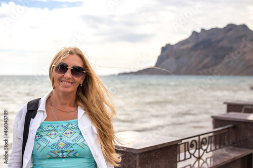 A blonde with long hair in sunglasses stands on the sea promenade and smiles. Travel and tourism. © Natalia