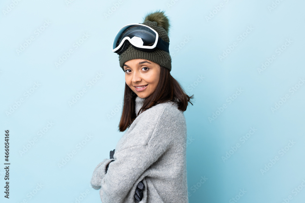 Mixed race skier girl with snowboarding glasses over isolated blue background with arms crossed and looking forward.