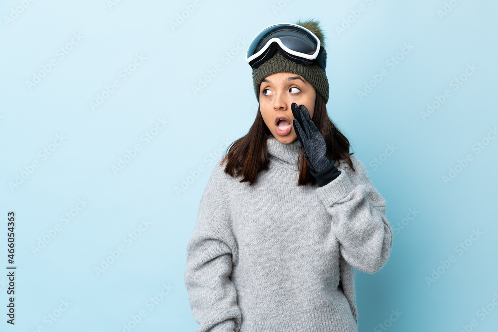 Mixed race skier girl with snowboarding glasses over isolated blue background whispering something with surprise gesture while looking to the side.