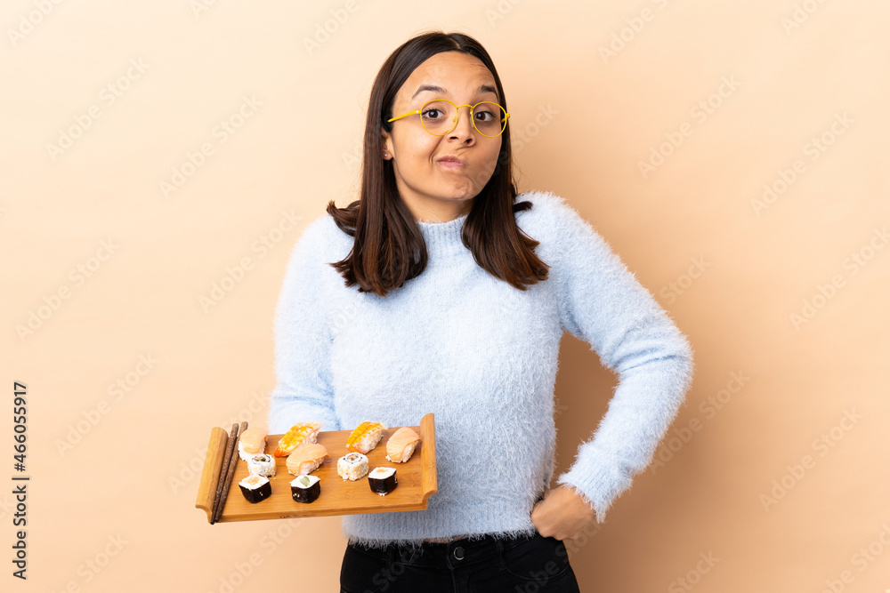 Young brunette mixed race woman holding sushi over isolated background making doubts gesture while lifting the shoulders Young brunette mixed race woman holding sushi over isolated background making doubts gesture while lifting the shoulders