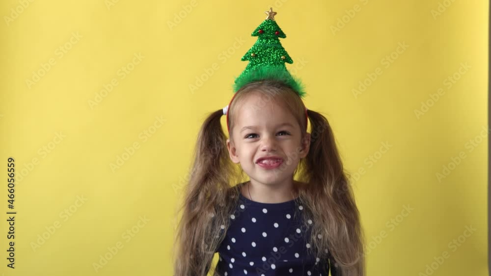 Portrait Emotion Cute Happy Preschool Baby Girl With Ponytail And Christmas Tree on Head Smiling Looking On Camera At Yellow Background. Child Celebrating Birthday. Kid Have Fun Spend New Year Time Portrait Emotion Cute Happy Preschool Baby Girl With Ponytail And Christmas Tree on Head Smiling Looking On Camera At Yellow Background. Child Celebrating Birthday. Kid Have Fun Spend New Year Time