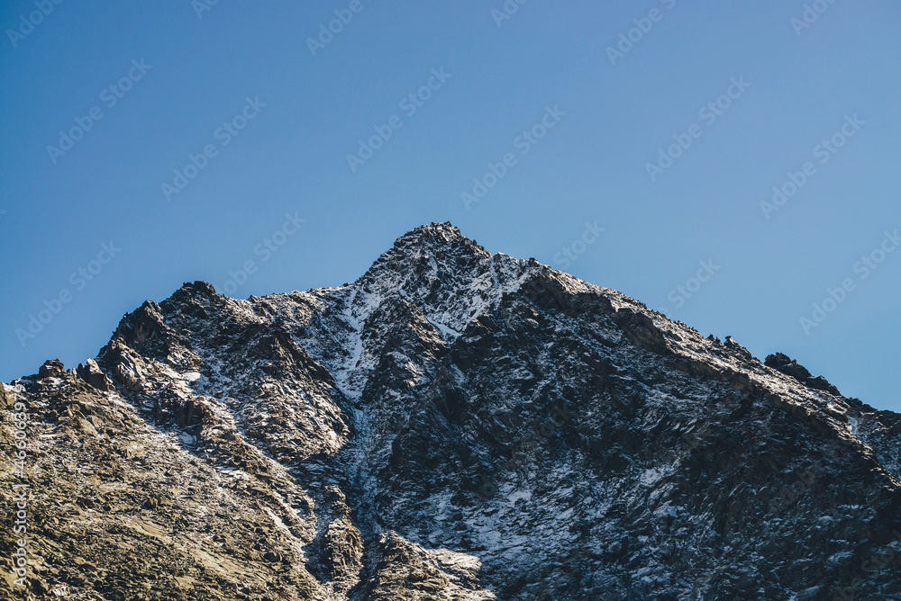 Awesome mountain landscape with black peaked top with white snow in golden sunshine under blue sky. Beautiful snow-covered pointy peak in gold sunlight. Atmospheric mountain scenery with peaked top.