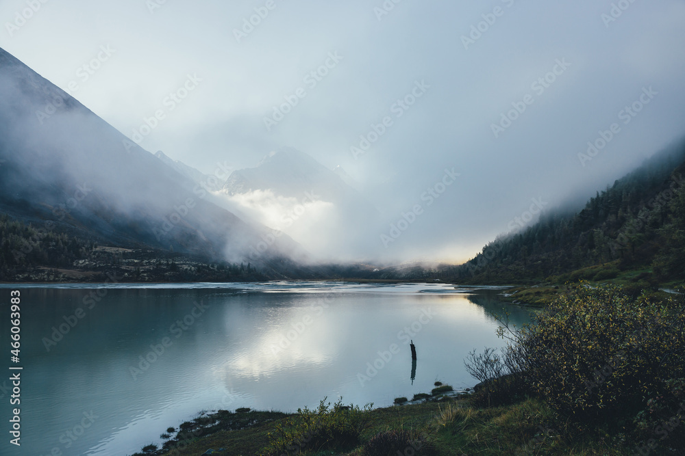 Atmospheric alpine landscape with mountain lake and high snow-covered mountain in dense low clouds. Beautiful low lighting scenery with pointy peak in thick fog and golden sunshine reflection in lake.