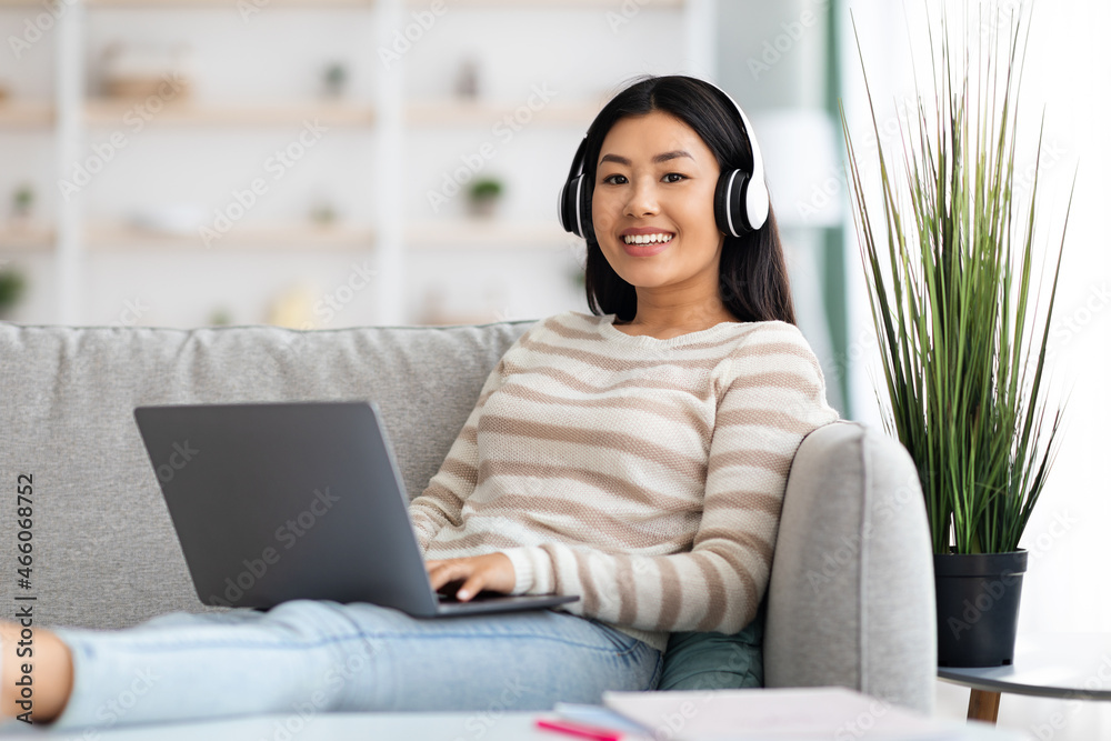 Naklejka premium Portrait Of Smiling Asian Lady Wearing Wireless Headphones Using Laptop At Home