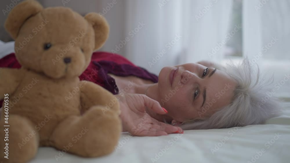 Side view happy woman lying on white bed smiling looking at camera with blurred teddy bear at front. Portrait of relaxed beautiful slim mature Caucasian lady posing in bedroom at home indoors Side view happy woman lying on white bed smiling looking at camera with blurred teddy bear at front. Portrait of relaxed beautiful slim mature Caucasian lady posing in bedroom at home indoors