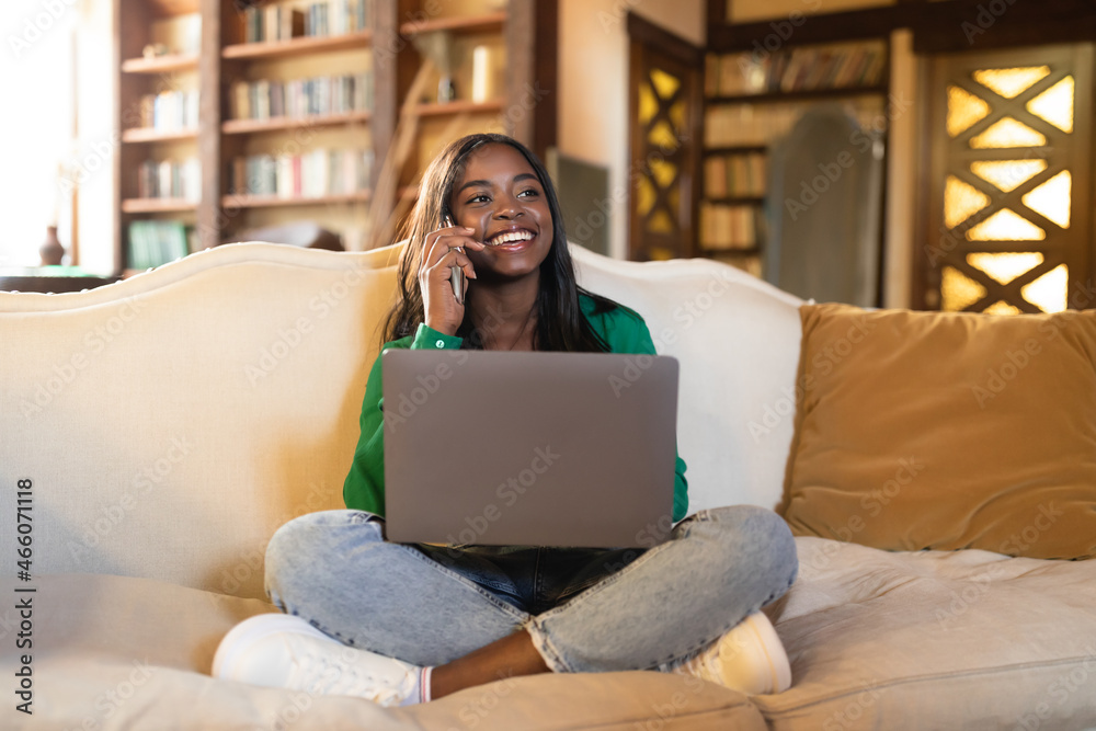Fototapeta premium Smiling young black woman having conversation on cellphone, working on laptop in at home