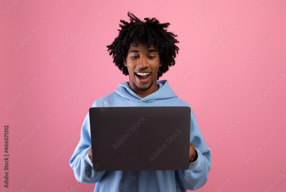 Happy black teenager looking at laptop screen with smile, watching interesting web content on pink studio background Happy black teenager looking at laptop screen with smile, watching interesting web content on pink studio background