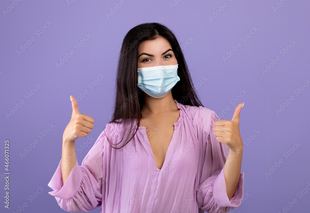 Portrait of armenian lady wearing medical face mask and gesturing thumbs up, posing to camera over purple background Portrait of armenian lady wearing medical face mask and gesturing thumbs up, posing to camera over purple background
