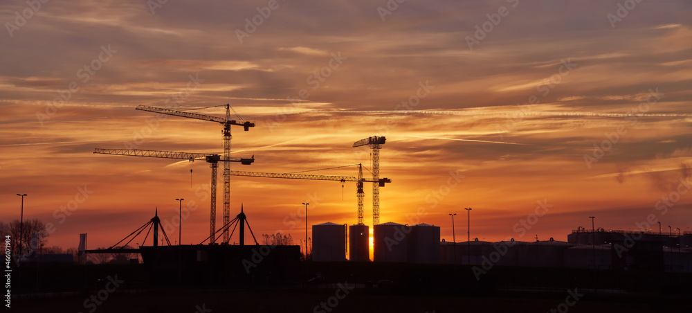 sunrise on a misty morning behind silhouettes of cranes and tanks in an industrial area