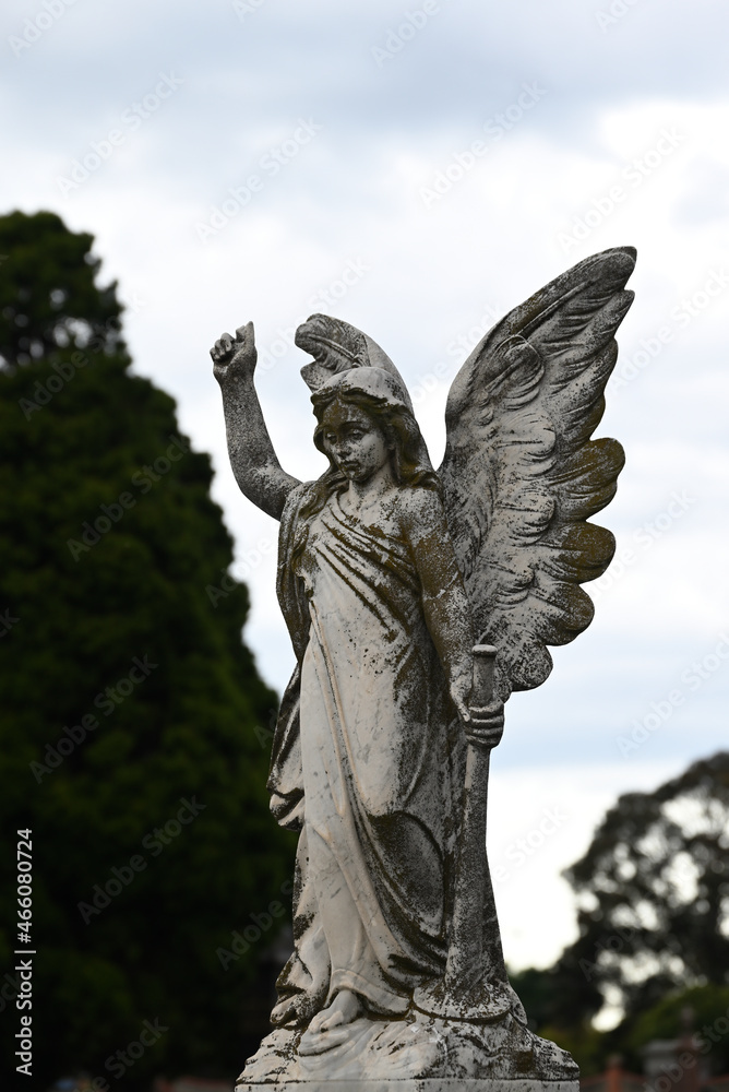 Fototapeta premium Stone sculpture of an angel holding a horn in a cemetery, with bright but cloudy sky in the background