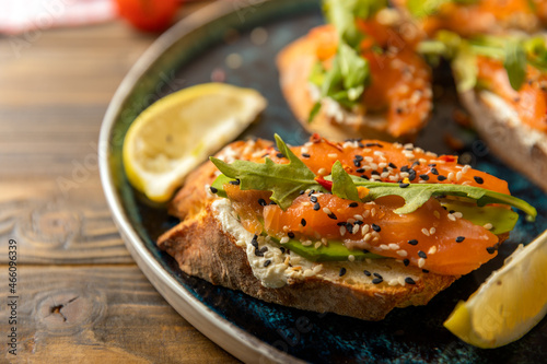 delicious bruschetta with salmon, avocado and arugula with lemon wedges on a large blue plate on a woody background.