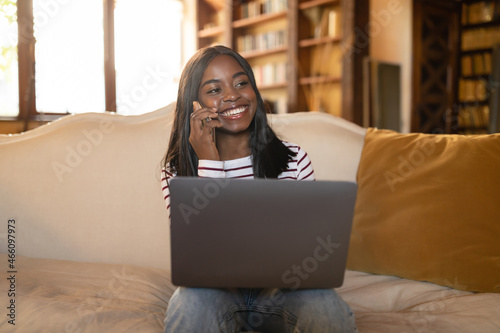 Online business meeting. Happy young black woman using laptop for video conference, calling on moile phone at home