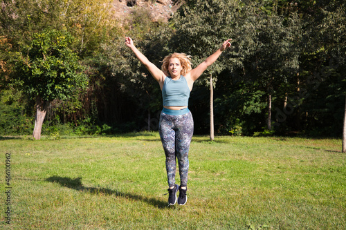 beautiful fat blonde woman playing sports in the park. She is jumping on the grass, spreading her arms and legs. Outdoor sport and calisthenics concept. Healthy life.