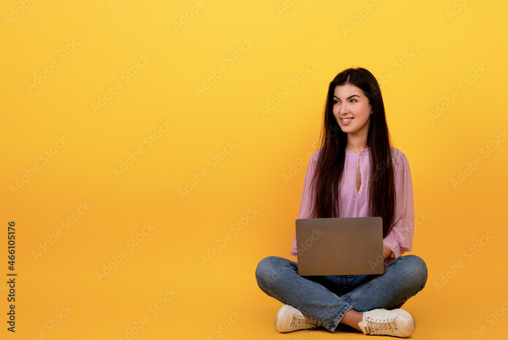 Happy armenian woman using laptop computer on yellow studio background, sitting on floor and looking at copy space Happy armenian woman using laptop computer on yellow studio background, sitting on floor and looking at copy space