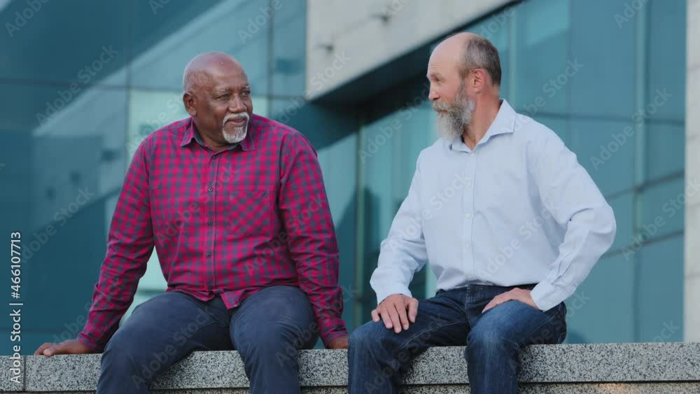 Two diverse bearded elderly male friends in casual clothes shirts and jeans sitting outdoors chatting carefree. Serious retirement age men, mature businessmen having conversation near office building Two diverse bearded elderly male friends in casual clothes shirts and jeans sitting outdoors chatting carefree. Serious retirement age men, mature businessmen having conversation near office building