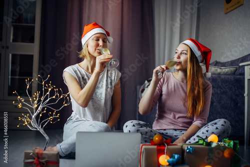 Two happy excited young women in Santa Claus Christmas hats drinking champagne sitting on the floor in a cozy home interior among gifts and burning garlands. © exebiche