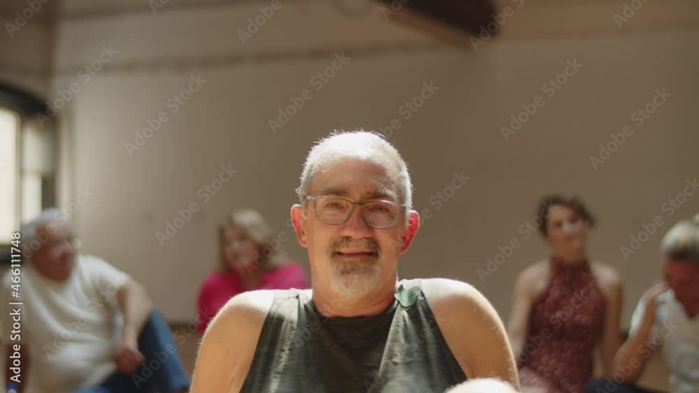 Slow motion of cheerful grey-haired senior man in dance studio. Medium shot of male mature person in sportswear sitting on floor, looking at camera, relaxing after training. Portrait, emotion concept Slow motion of cheerful grey-haired senior man in dance studio. Medium shot of male mature person in sportswear sitting on floor, looking at camera, relaxing after training. Portrait, emotion concept