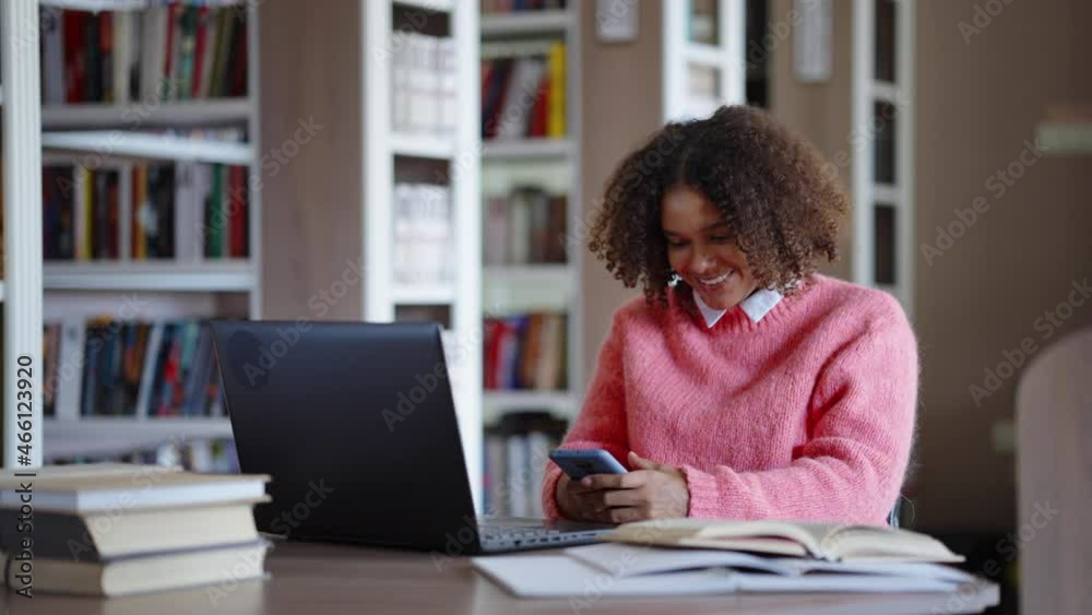 Black girl with curly hair wearing pink sweater sitting in library and texting message on smartphone, laptop and books in front of her, bookcases on blurred background. Student communicating online Black girl with curly hair wearing pink sweater sitting in library and texting message on smartphone, laptop and books in front of her, bookcases on blurred background. Student communicating online