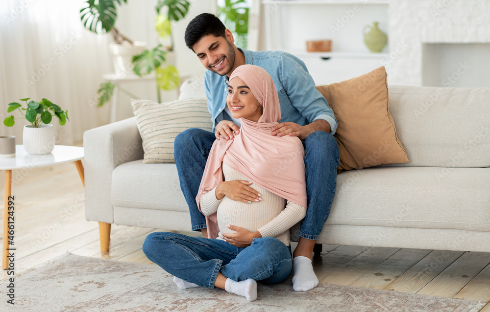 Pregnant muslim woman feeling relaxed, sitting on floor at living room and attentive husband massaging her shoulders