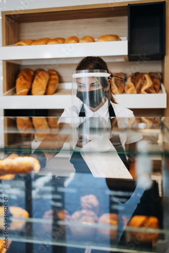 Beautiful young female worker with protective mask and face shield working in bakery. Business and coronavirus pandemic concept.