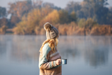 Woman relaxing with coffee next to lake at autumn cold morning. Camping and hiking outdoors at fall season. Blond hair woman wearing knit hat and sweater