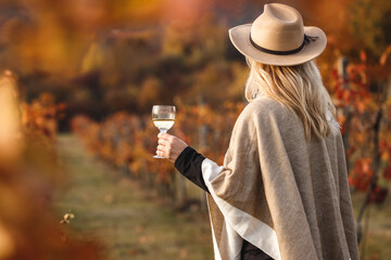 Female vintner enjoying white wine in her vineyard. Woman with poncho and hat holding glass of wine outdoors. Winery at autumn
