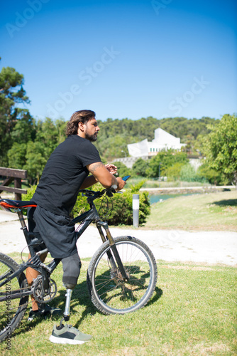 Side view of man with mechanical leg on bicycle. Thoughtful person with disability sitting on bicycle, phone in hands. Sport, disability concept
