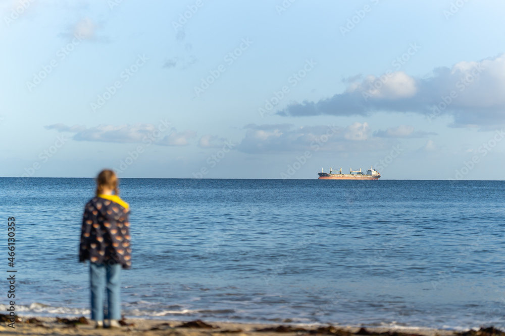 Young girl out of focus stood on beach looking out to sea in sunshine at big ocean liner oil tanker on the horizon. Enormous oil tanker delivering fuel and petrol to England. 