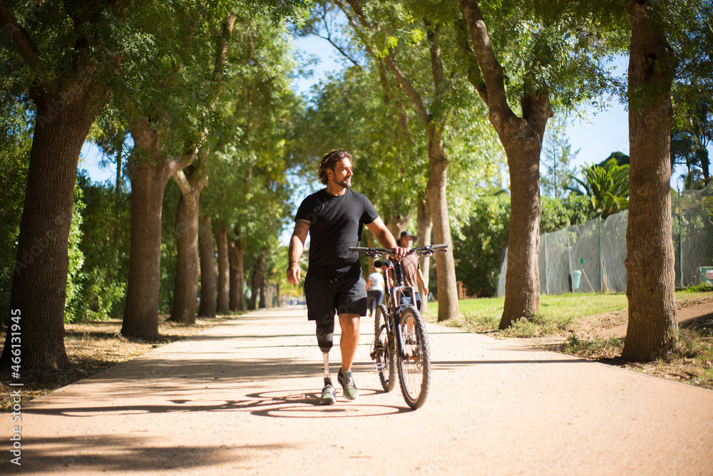 Front view of person with disability with bicycle. Curly man with mechanical leg walking in park on sunny day. Sport, disability concept