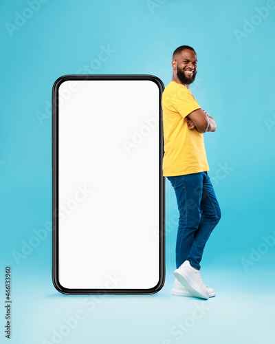 Hot offer, online sale. Funky black young guy standing near huge cellphone with mockup space over blue background