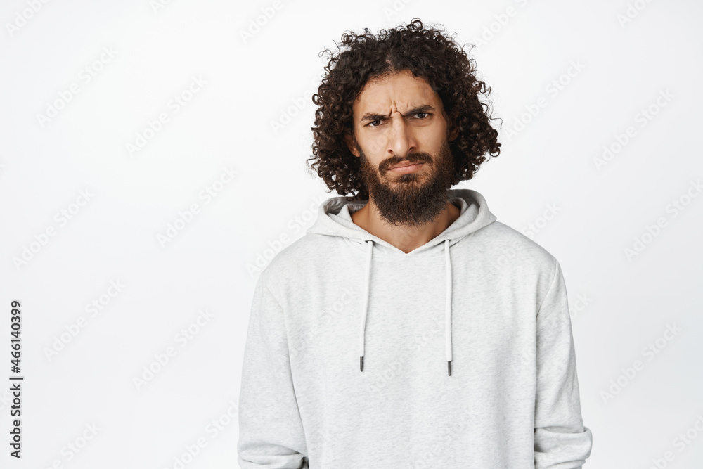 Portrait of moody young man sulking, furrow eyebrows and looking disappointed, standing in hoodie over white background Portrait of moody young man sulking, furrow eyebrows and looking disappointed, standing in hoodie over white background
