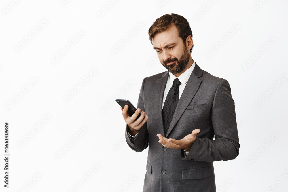 Image of displeased businessman looking at mobile phone app screen and grimacing slightly disappointed, checking applications stats, white background Image of displeased businessman looking at mobile phone app screen and grimacing slightly disappointed, checking applications stats, white background