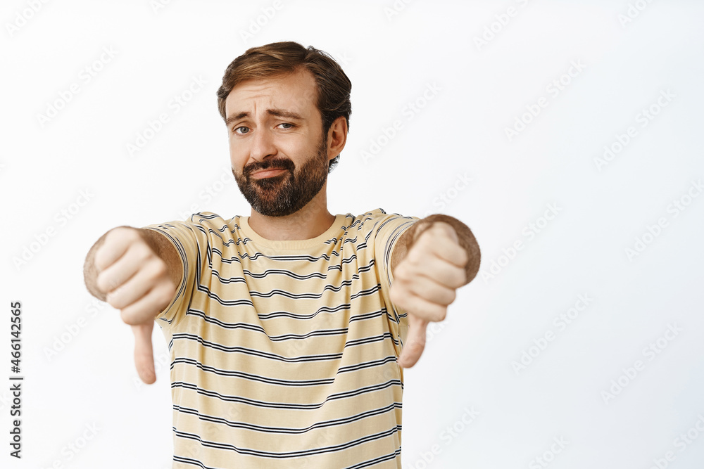 Fototapeta premium Very bad. Portrait of bearded guy shows thumbs down and smirks with dislike, disappointed by smth, standing over white background