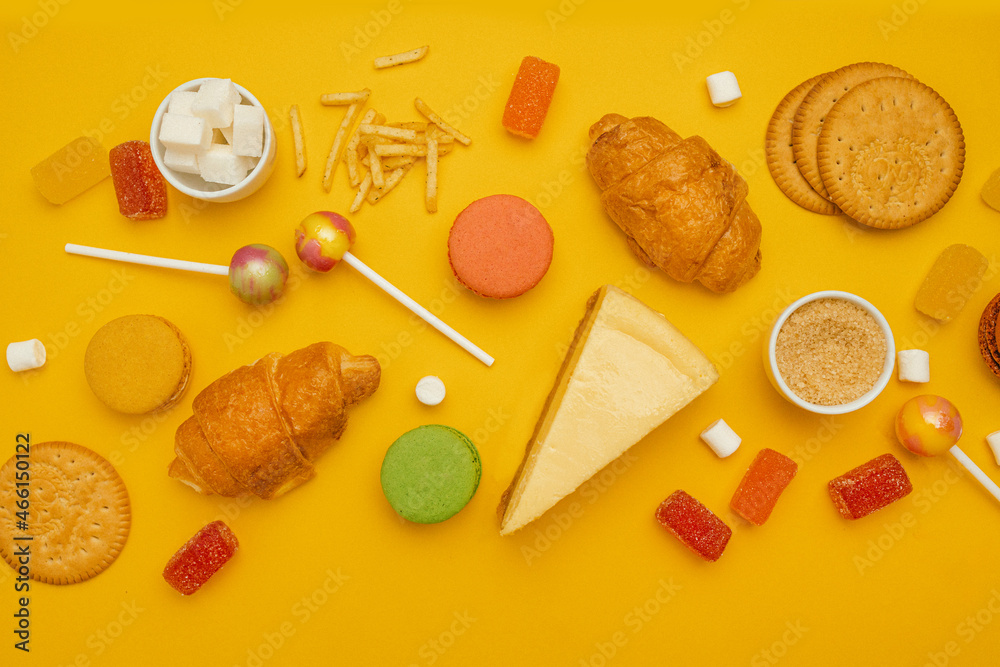 Unhealthy foods on a colored background, top view. French croissant, candy on a stick, jelly candies, marshmallows, cookies and macarons on a yellow bright background Unhealthy foods on a colored background, top view. French croissant, candy on a stick, jelly candies, marshmallows, cookies and macarons on a yellow bright background
