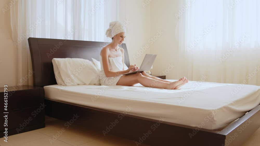 Caucasian woman using wireless laptop while resting on bed in bath towel. Female traveler enjoying time at comfy hotel room with modern device. Woman in bath towel relaxing at hotel room with laptop. Caucasian woman using wireless laptop while resting on bed in bath towel. Female traveler enjoying time at comfy hotel room with modern device. Woman in bath towel relaxing at hotel room with laptop.