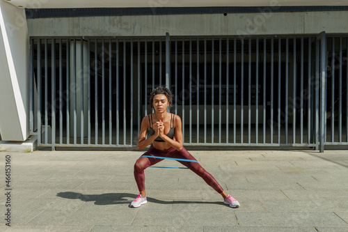 Attractive young woman in stylish tracksuit does side squats with elastic band and keeps hands on waist near modern building on sunny city street