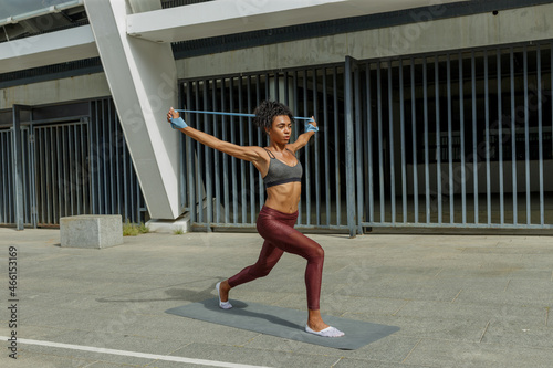 Attractive sportswoman in tracksuit does dynamic lunges raising hands with elastic band near modern building on sunny street © sergiymolchenko