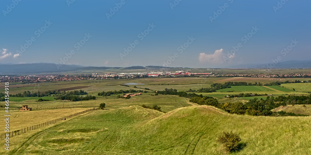 Transylvanian landscape with green fields with and a city and mountains in the distance on a sunny day with clear blue sky, view from above ffrom Rapa Rosie, Romania 