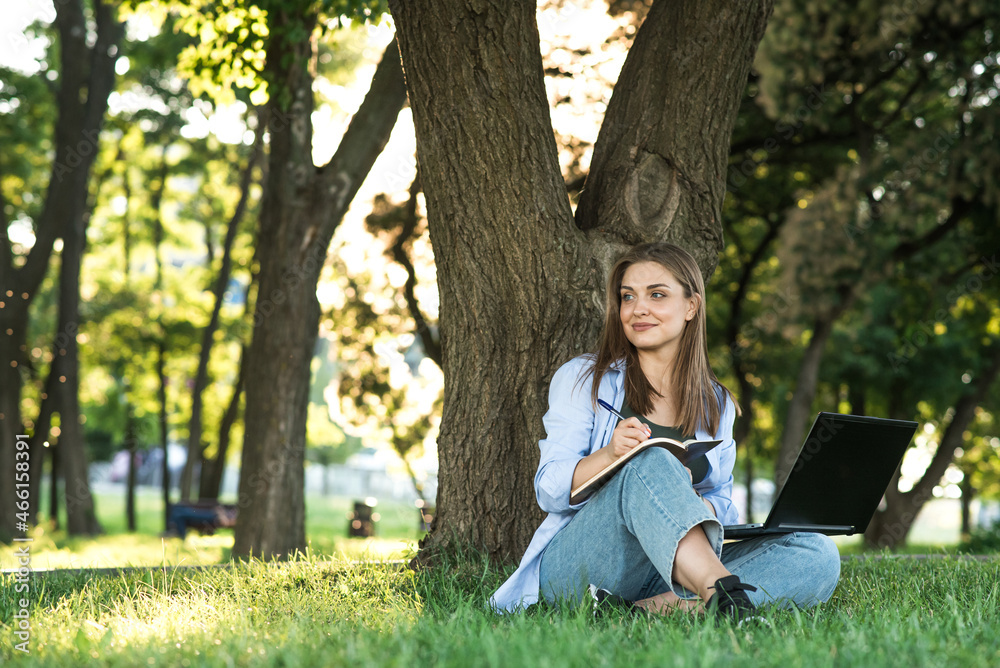 A young and attractive Caucasian girl sitting on the grass on the lawn and working. Writing in a notebook. Freelance and education concept A young and attractive Caucasian girl sitting on the grass on the lawn and working. Writing in a notebook. Freelance and education concept