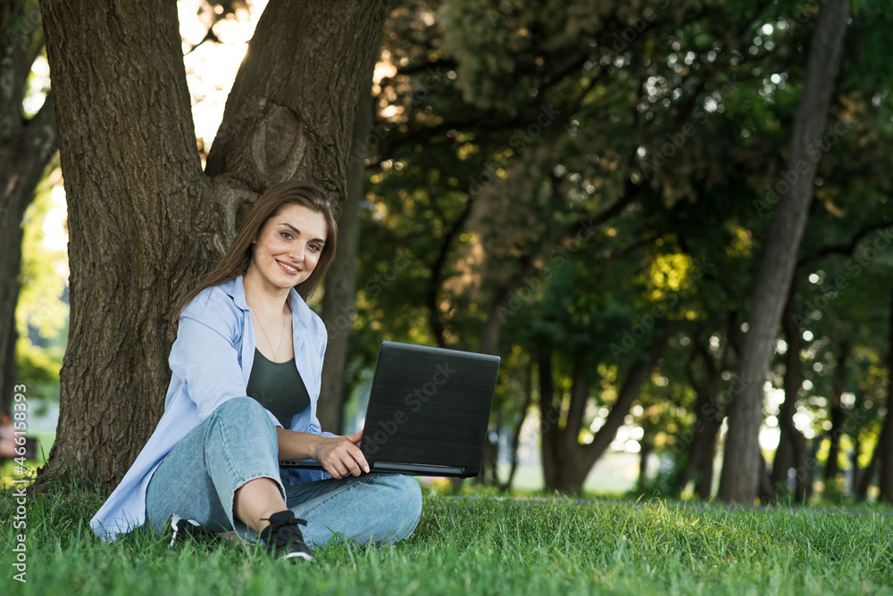 A young and attractive Caucasian girl sitting on the grass on the lawn and working. Using a laptop. Freelance and education concept. A young and attractive Caucasian girl sitting on the grass on the lawn and working. Using a laptop. Freelance and education concept.