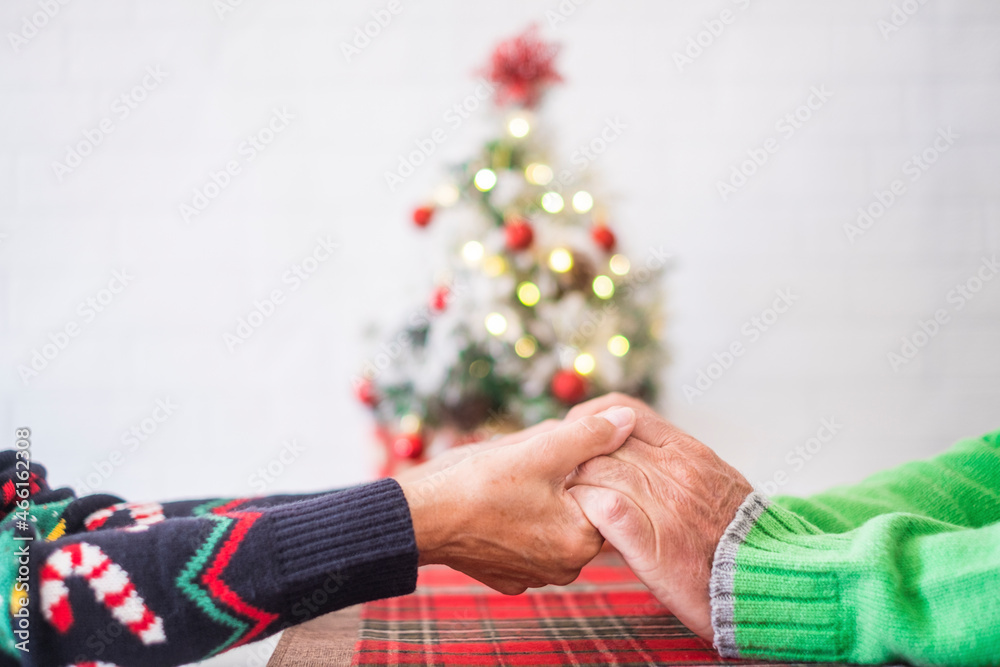 Senior couple in warm clothing holding each others hands in front of decorated christmas tree at home. Loving old romantic heterosexual couple celebrating christmas festival together