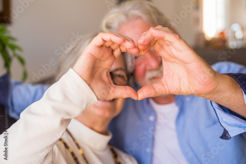 Couple of two old and happy seniors having fun at home on the sofa doing a heart shape with their hands and fingers looking at the camera. .