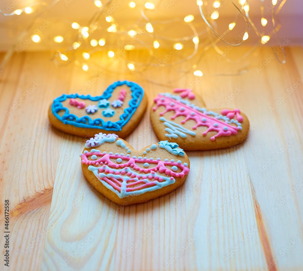 Fresh baked appetizing beautiful tasty glazed gingerbread cookies shape of heart with colored pattern, brown wooden table on background of New Year garland thread with light bulbs warm yellow color