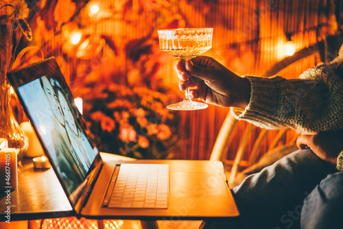 Young woman takes online congratulations for holiday via laptop holding wineglass with drink sitting on balcony decorated with lit candles and lights