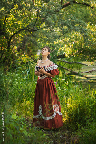 Sad young woman in a 19th century dress by the river with a book in her hands. Summer landscape. The photo.