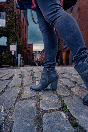 A women wearing jeans and black boots walks down a cobblestone street.