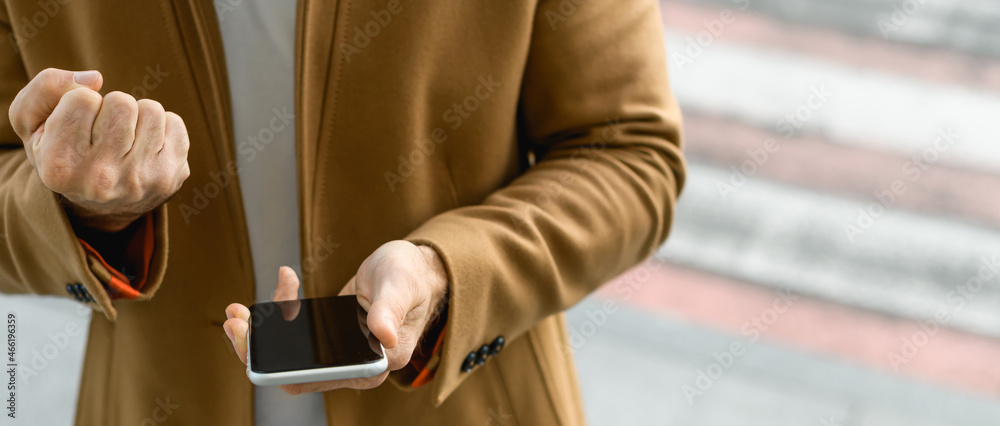 Cropped view of man in coat holding smartphone and showing yes gesture outdoors, panorama